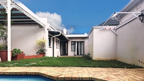 A swimming pool with a tiled edge, a grassy yard, and a white building with a covered patio area. - Selton Guest House (Cape Town)