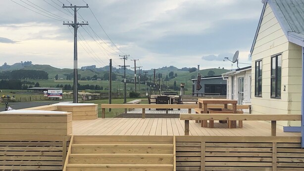 A wooden deck with benches and a picnic table, overlooking a rural landscape with hills and a house with a satellite dish. - Atiamuri Bach Family (Atiamuri)