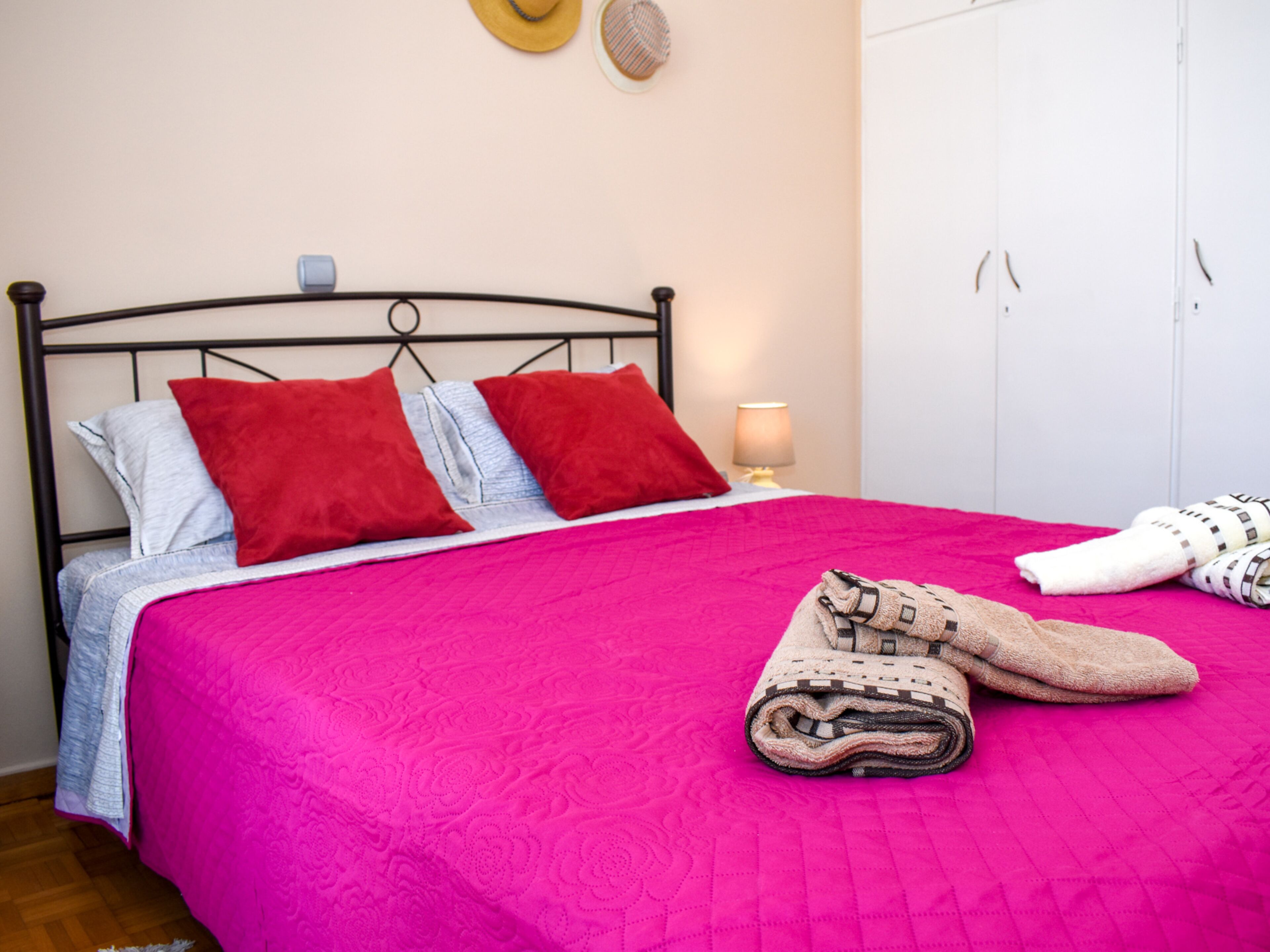 Una cama bien hecha con una colcha rosa brillante, dos almohadas rojas y una toalla doblada al lado.