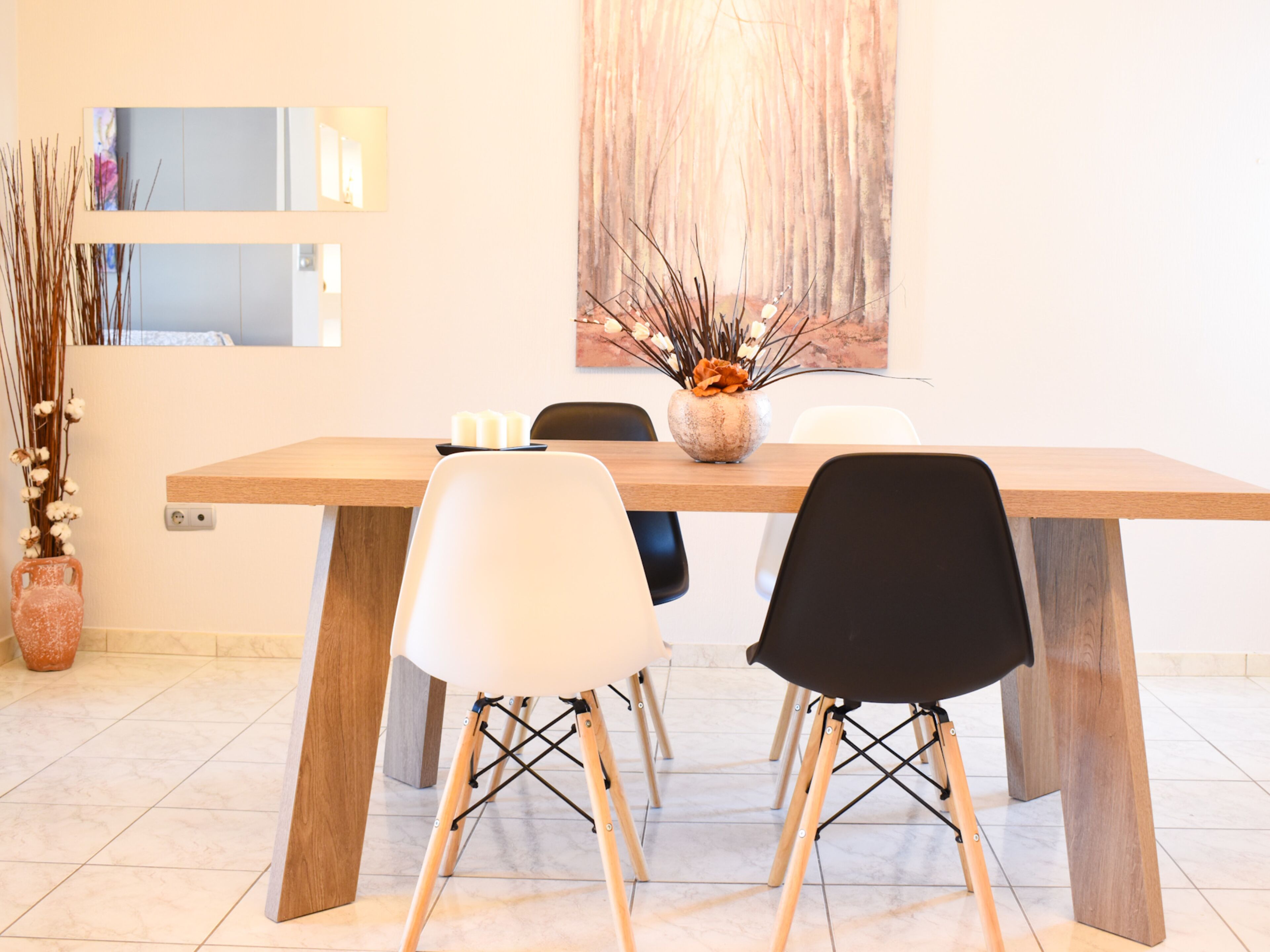 A dining area with a wooden table, four chairs, a vase with dried plants, and a large abstract painting on the wall.