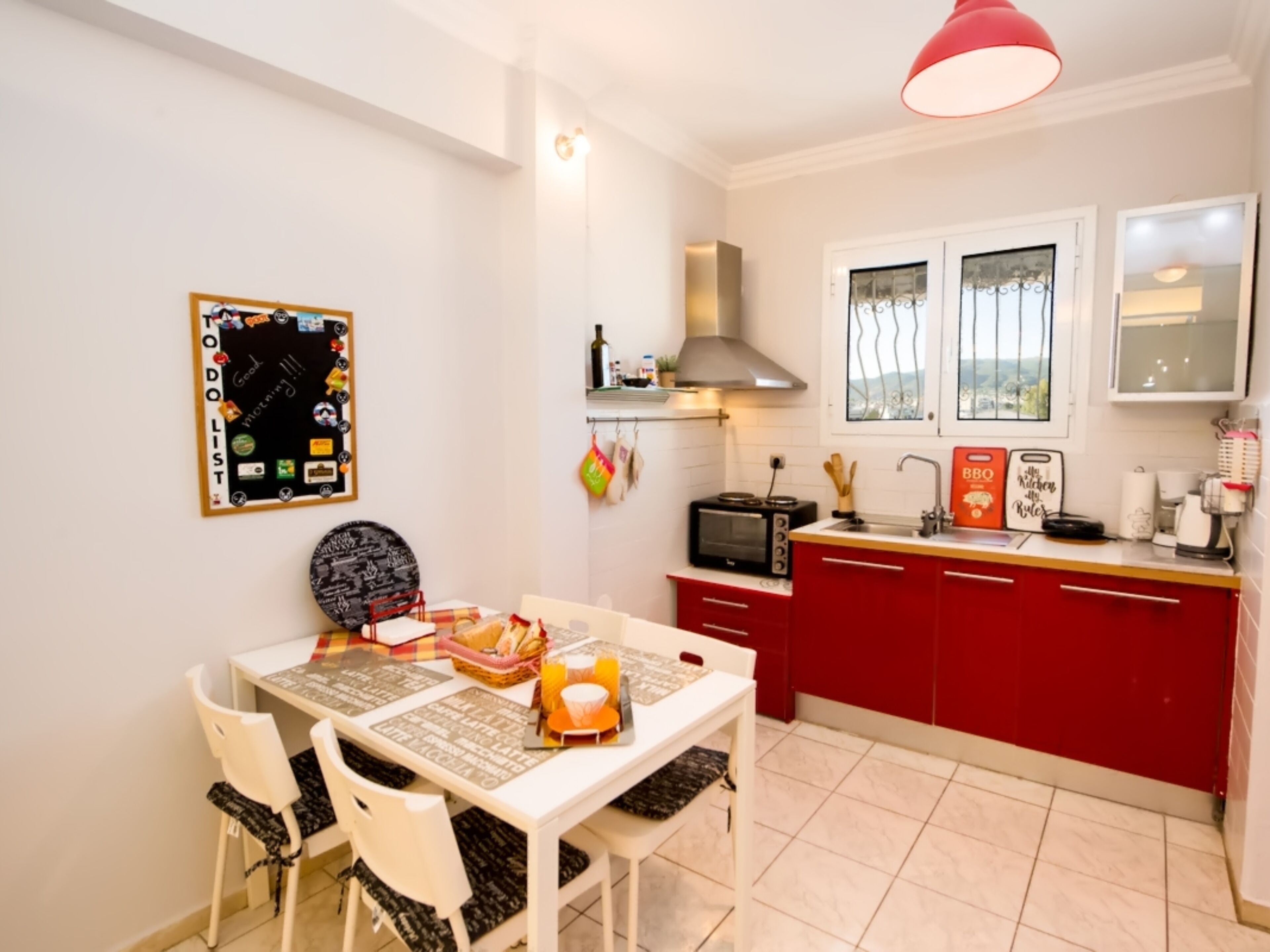 A compact kitchen with a red cabinet, a stove, and a microwave. There is a dining table with chairs and a basket of bread.