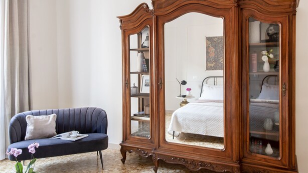 Ein Schlafzimmer mit einem blauen Sofa, einem Holzschrank mit Glastüren, einem Bett mit weißer Bettwäsche und einem kleinen Tisch mit einer Vase.