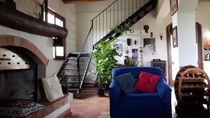 A cozy living room with a fireplace, a blue armchair, a staircase, and various decorations. - Affittacamere Luti (Oristano)