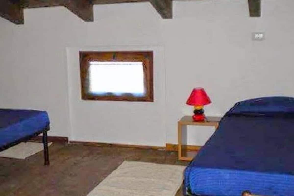 A room with two beds, a small window, a red lamp on a nightstand, and a wooden floor.
