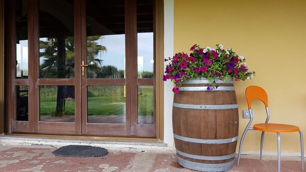 A wooden barrel planter with flowers next to a chair, a door with glass panels, and a doormat on a porch. - Affittacamere Luti (Oristano)