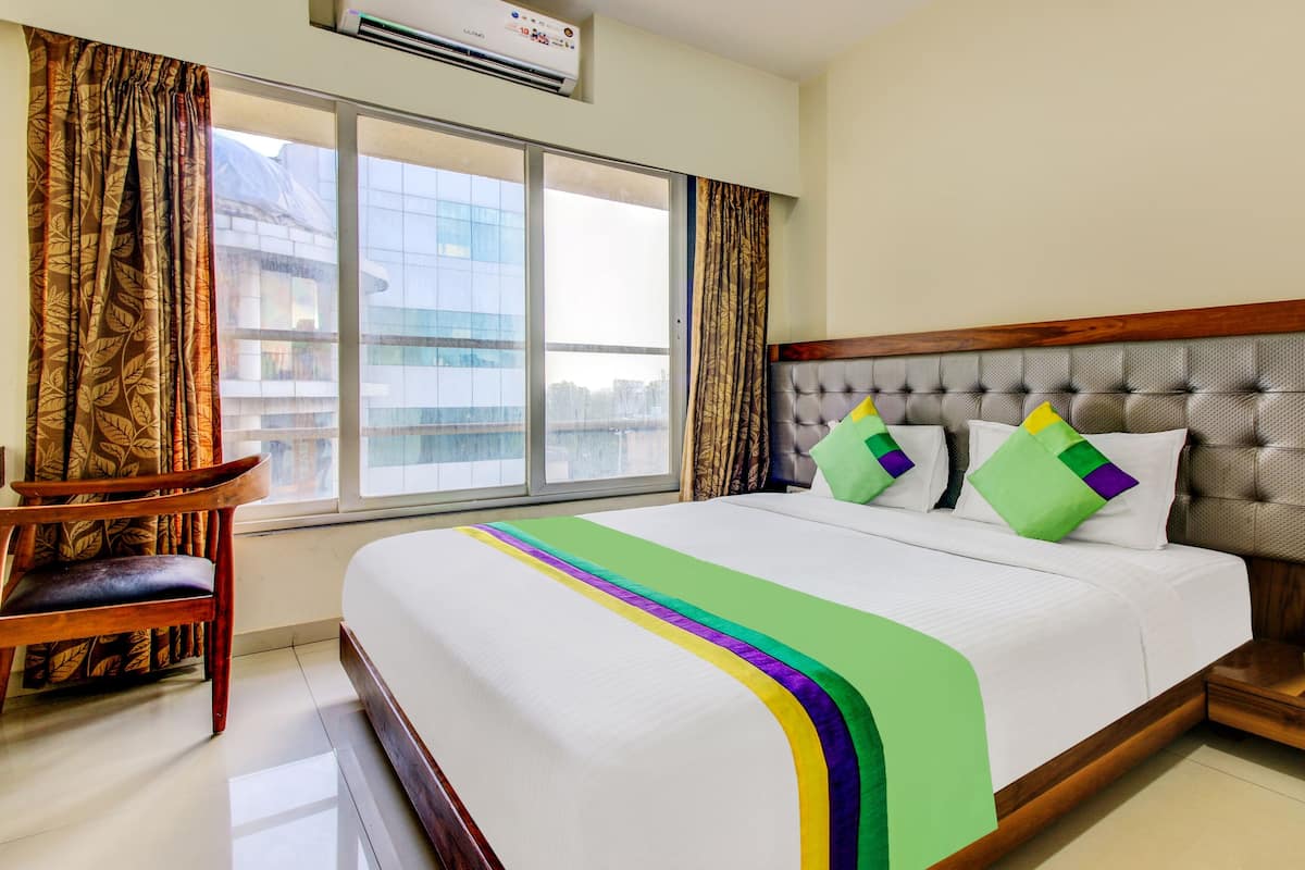 Una habitación de hotel con una cama grande, una mesita de noche de madera, una silla y un ventanal con cortinas.