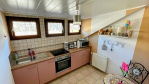 A compact kitchen with a sink, stove, and microwave. There are two windows with curtains, a radiator, and a small dining table with a chair. - Cozy vacation apartment "Schwalbennest" under the roof with garden access (Bundenthal)
