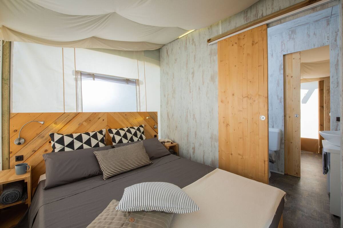 Ein modernes Schlafzimmer mit einem Holz-Kopfteil, einem großen Bett mit vielen Kissen, einem Nachttisch mit Lampe und Blick ins Badezimmer durch eine geöffnete Tür.