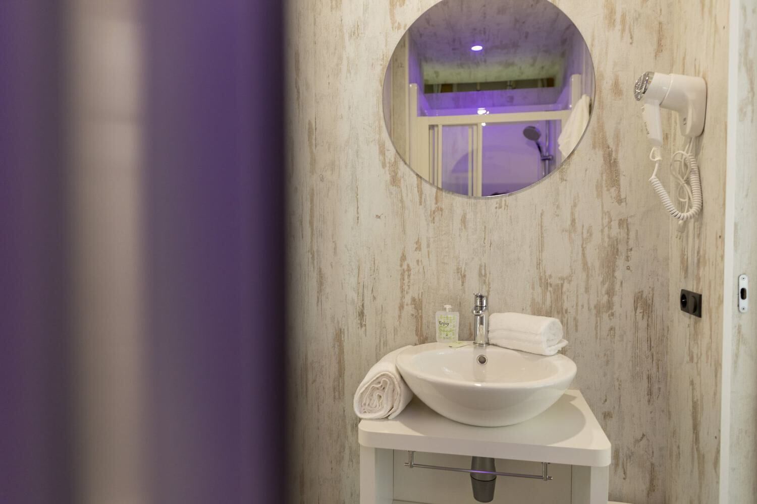Ein Badezimmer mit rundem Spiegel, Waschbecken in Weiß, einem Föhn und einem Handtuch.