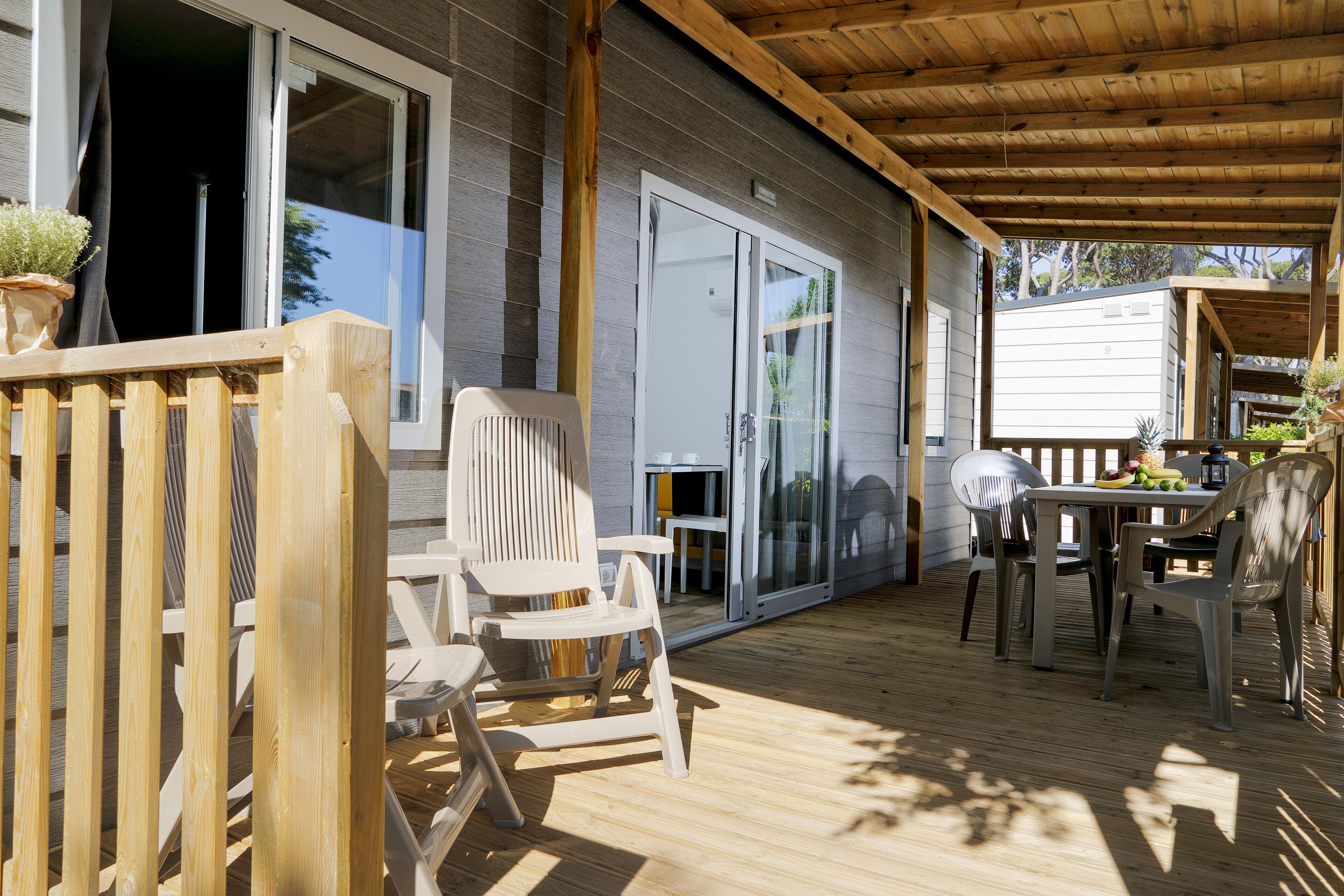 Una terraza de madera con mobiliario exterior, una puerta corrediza de vidrio y una zona cubierta con una mesa y sillas.