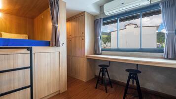 A compact room with a bunk bed, a desk with two stools, a window with curtains, and an air conditioning unit.