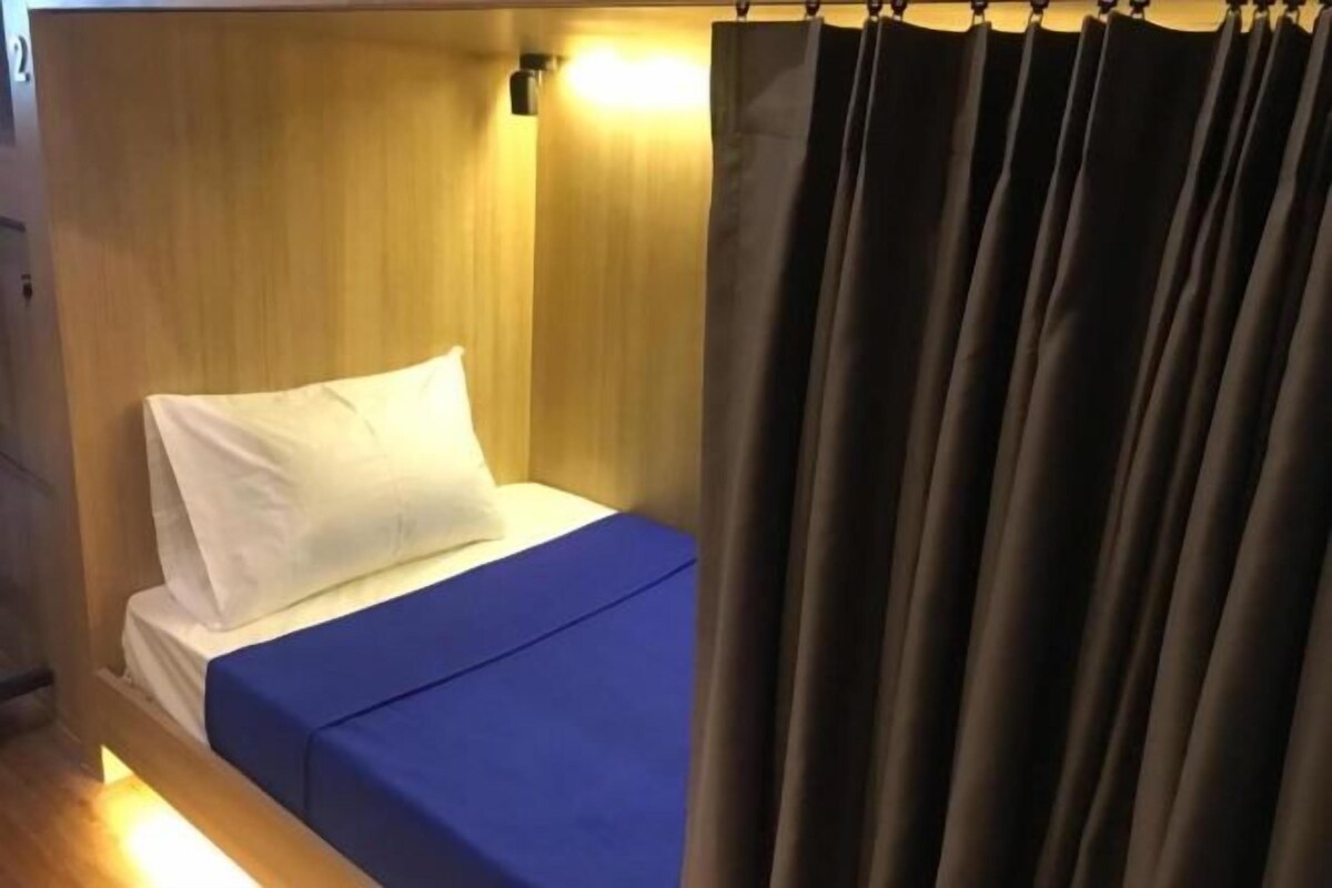 A single bed with a white pillow and a blue bedspread in a room with wooden paneling and a curtain.