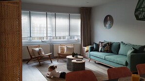 Un salon moderne comprenant un canapé, des fauteuils, une table à manger et une grande fenêtre équipée de stores.