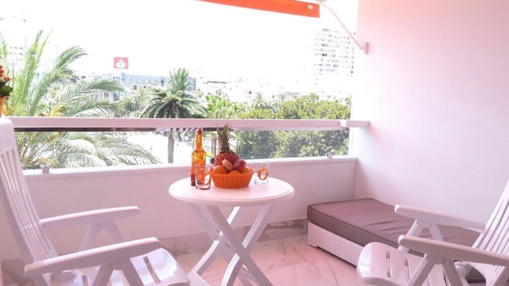 A balcony with a white table, two white chairs, a wicker sofa, and a view of palm trees and buildings.