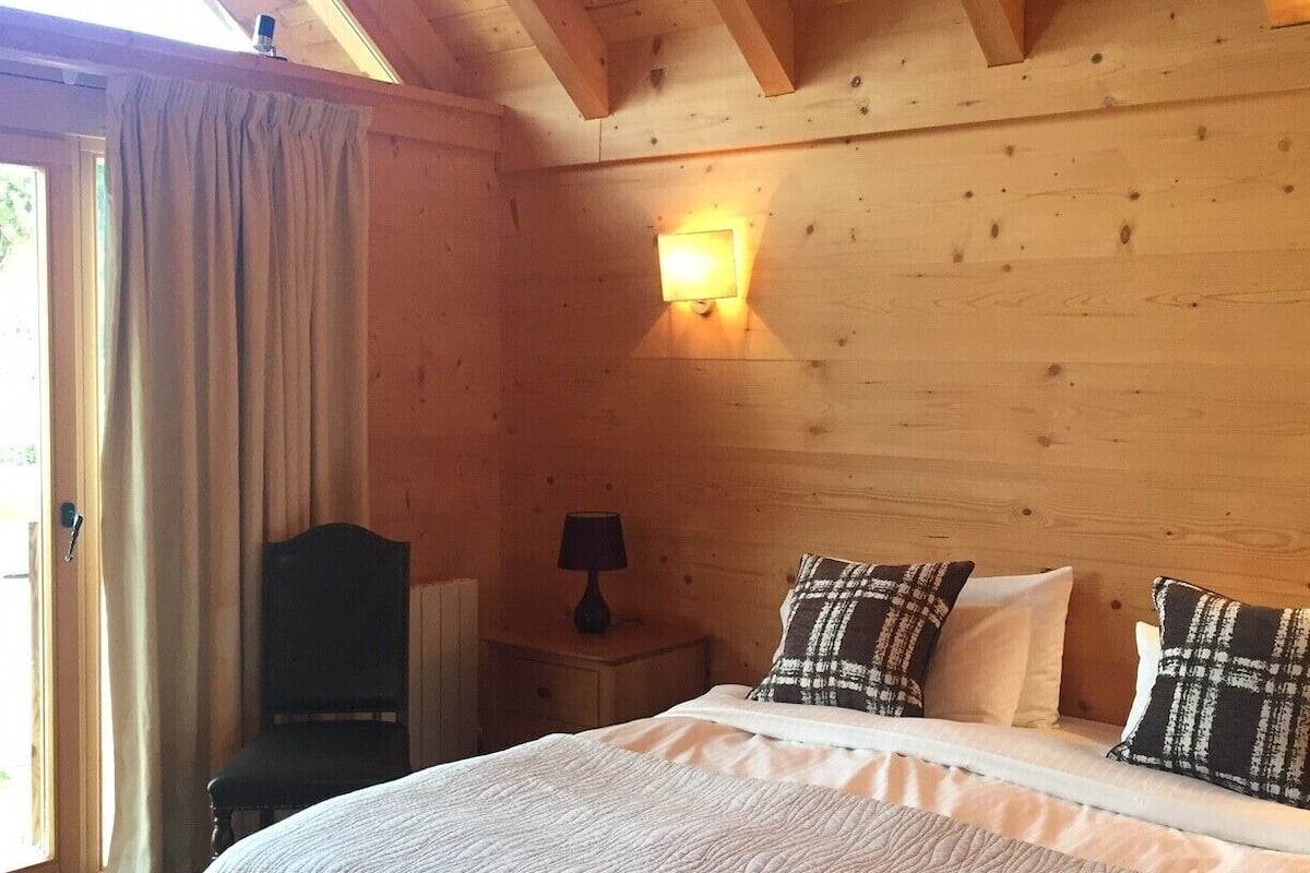 A wooden cabin room with a bed, a nightstand, a lamp, and a window with curtains.