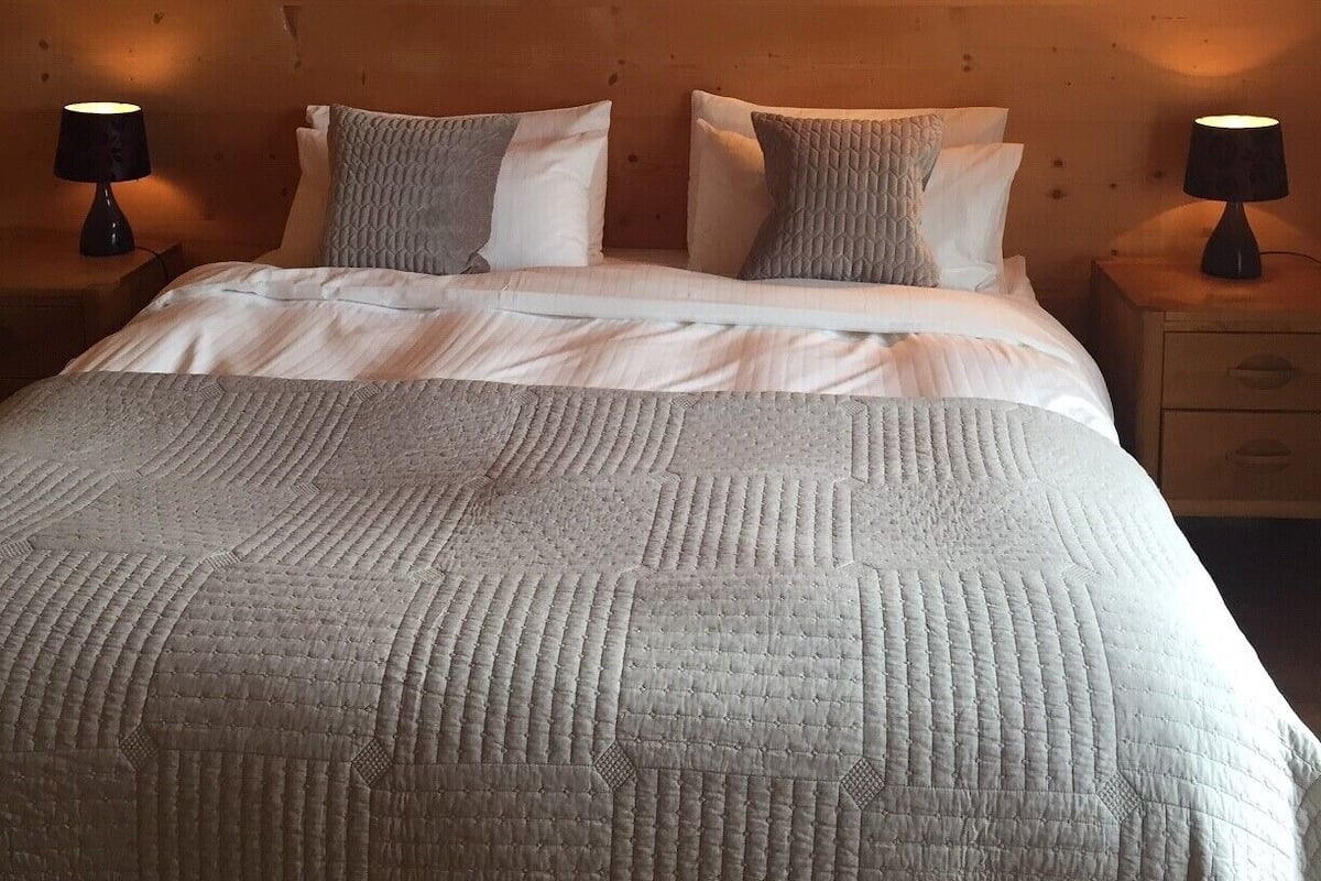 A cozy bedroom with a wooden ceiling, a large bed with a textured blanket, two bedside lamps, and a wall-mounted light fixture.