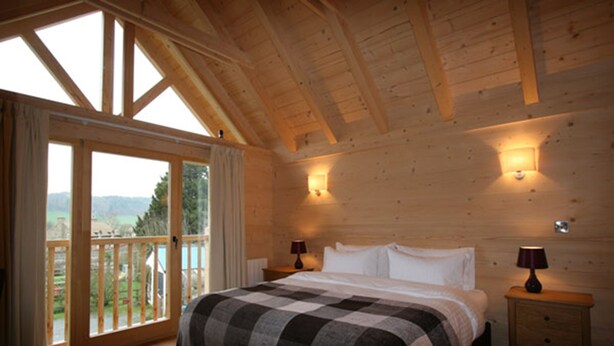 A wooden cabin bedroom with a large bed, two bedside tables, and a balcony with a view of the outdoors. - Pandy B&B (Hereford)