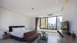 A modern hotel room with a large bed, a sofa, a glass coffee table, a TV, and a city view.