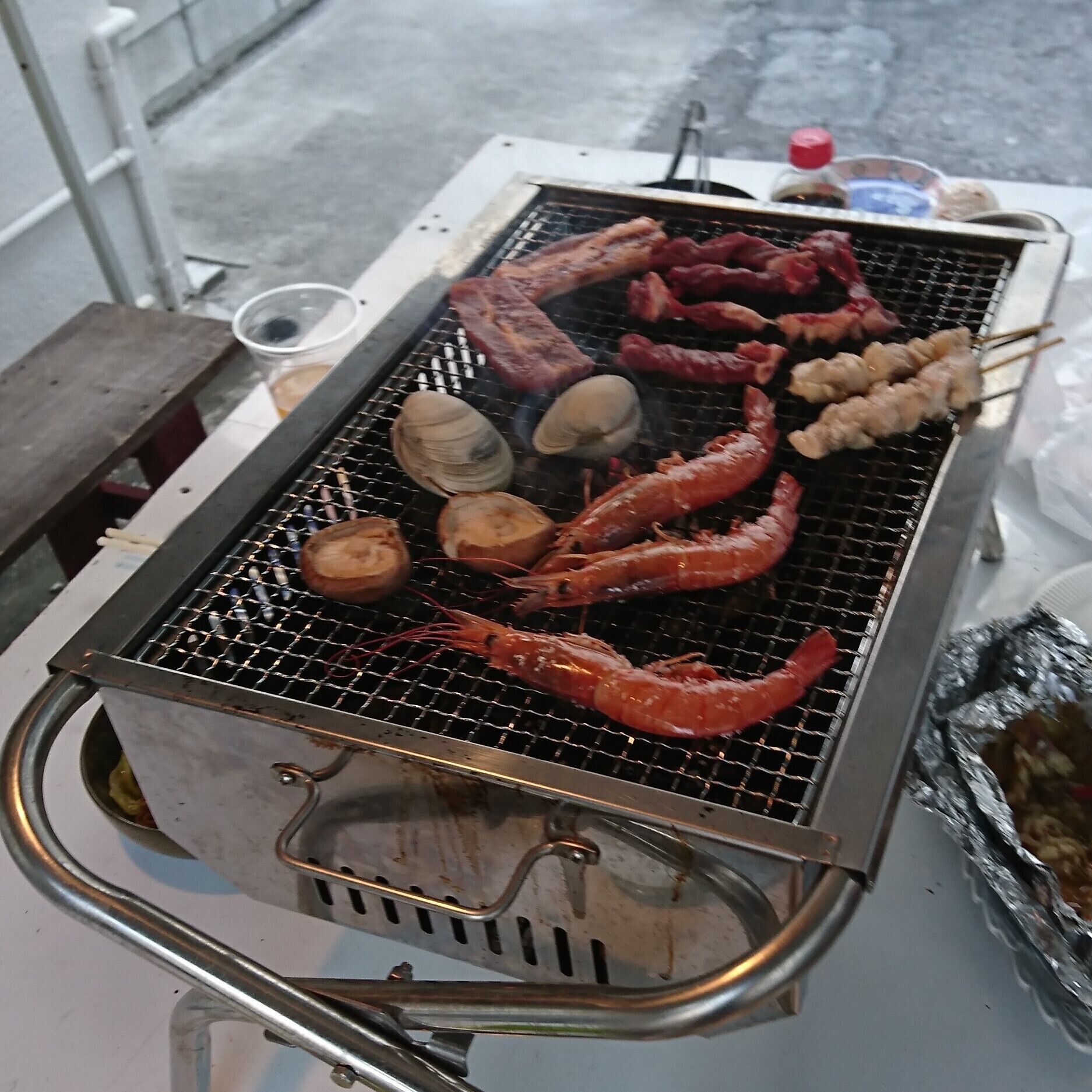 A portable grill with various meats and seafood being cooked.