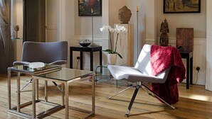 A modern living room with a glass coffee table, a white armchair with a red throw, and a grey armchair. There are two paintings on the wall, a Buddha statue, and a potted plant. - Maison de Myon (Nancy)