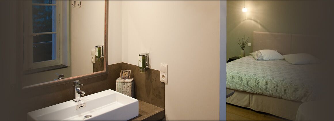 A bathroom with a sink, mirror, and window. The room has a bed with a headboard and a bedside table.