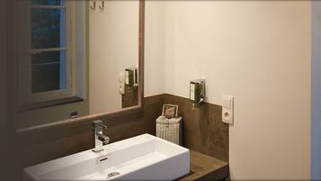 A bathroom with a sink, mirror, and window. The room has a bed with a headboard and a bedside table.