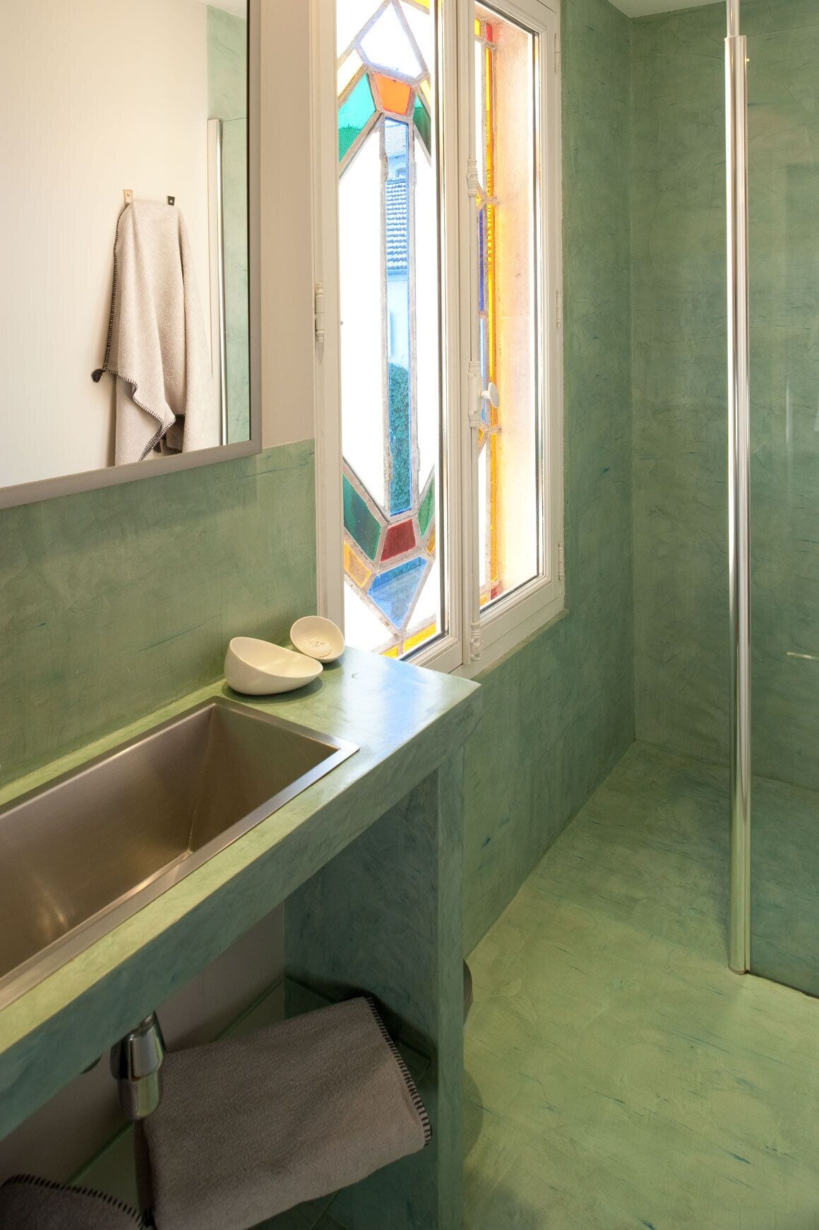 Un baño con pared de azulejos verdes, un lavamanos de acero inoxidable, cabina de ducha de vidrio y una ventana con vitral.