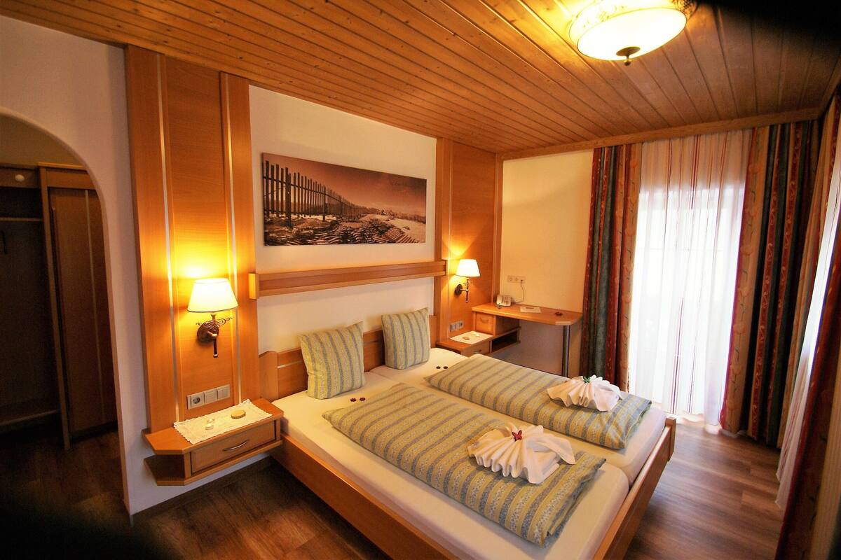 Een hotelkamer met een houten plafond, twee bedden met gestreept beddengoed, nachtlampjes, een schilderij aan de muur en een raam met gordijnen.