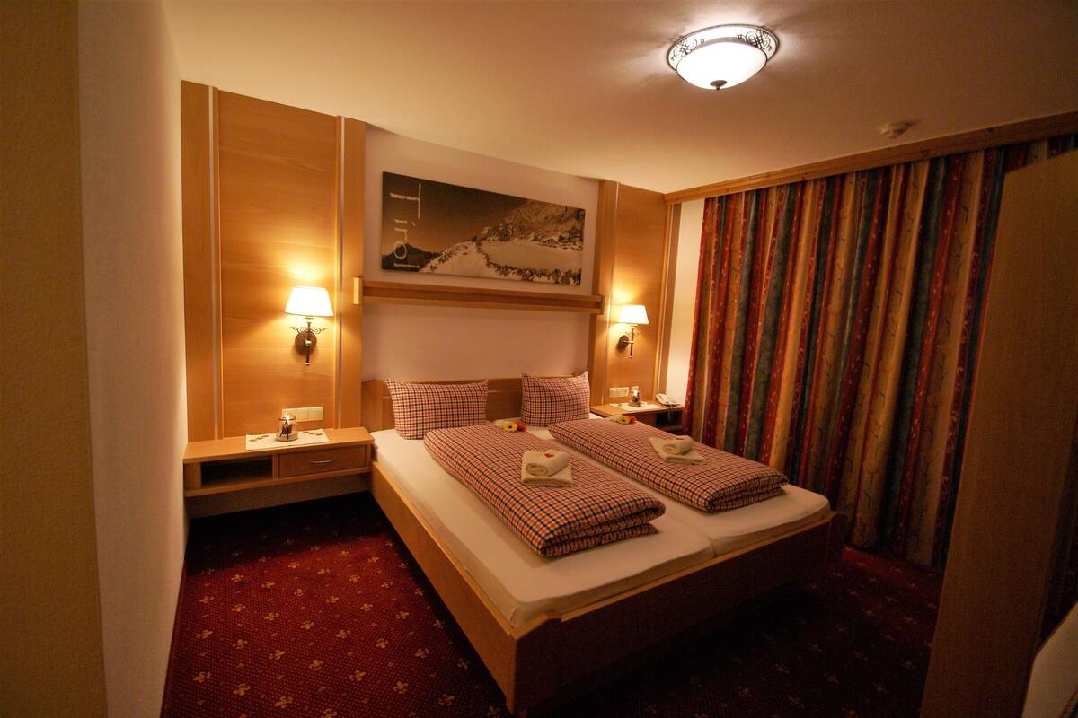 Een hotelkamer met een tweepersoonsbed, twee nachtkastjes met lampen, een hoofdbord met een schilderij en een gordijn met een patroon.