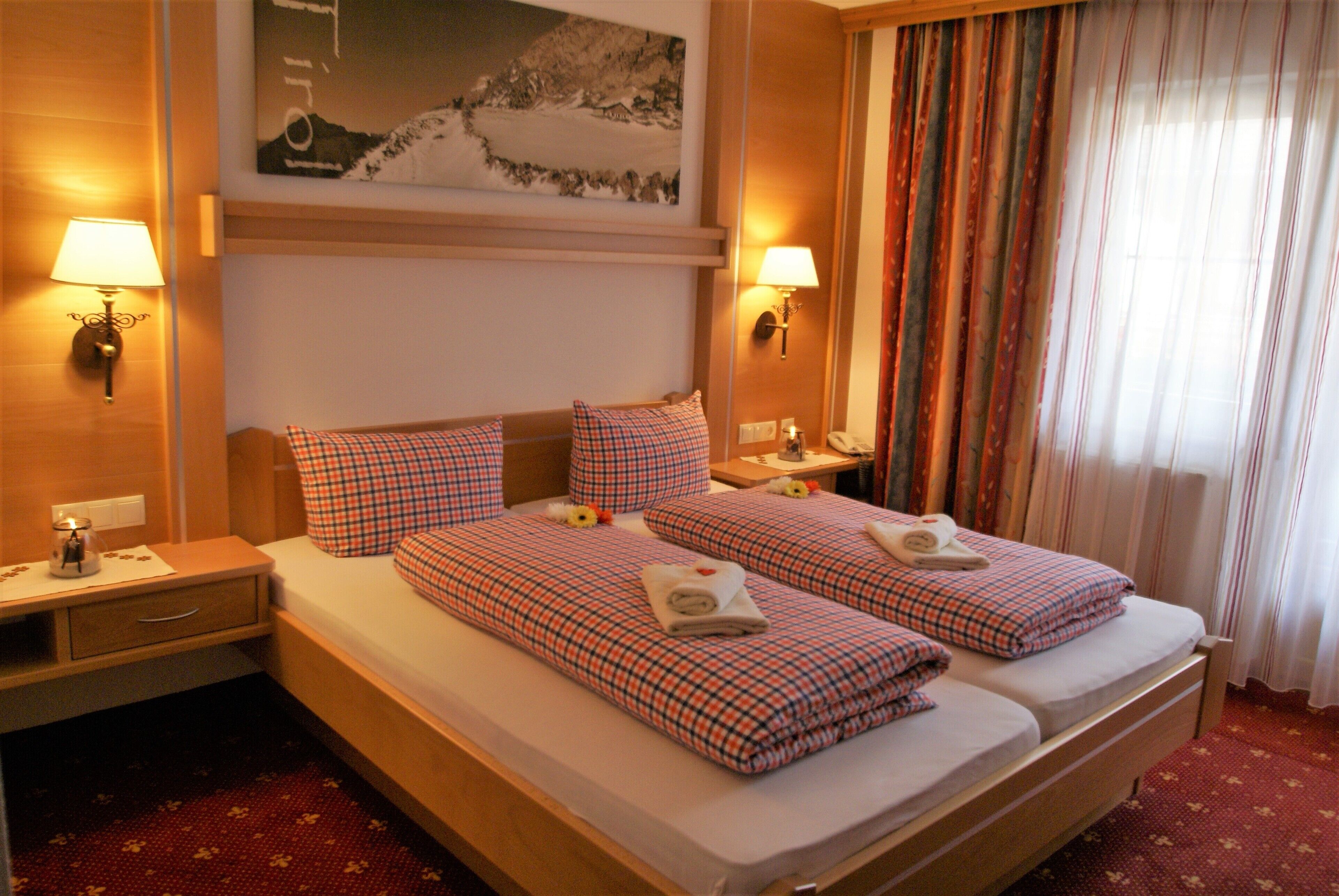 A hotel room with two beds, each with a checkered bedspread and pillows, a nightstand with a lamp, a painting on the wall, and a window with curtains.