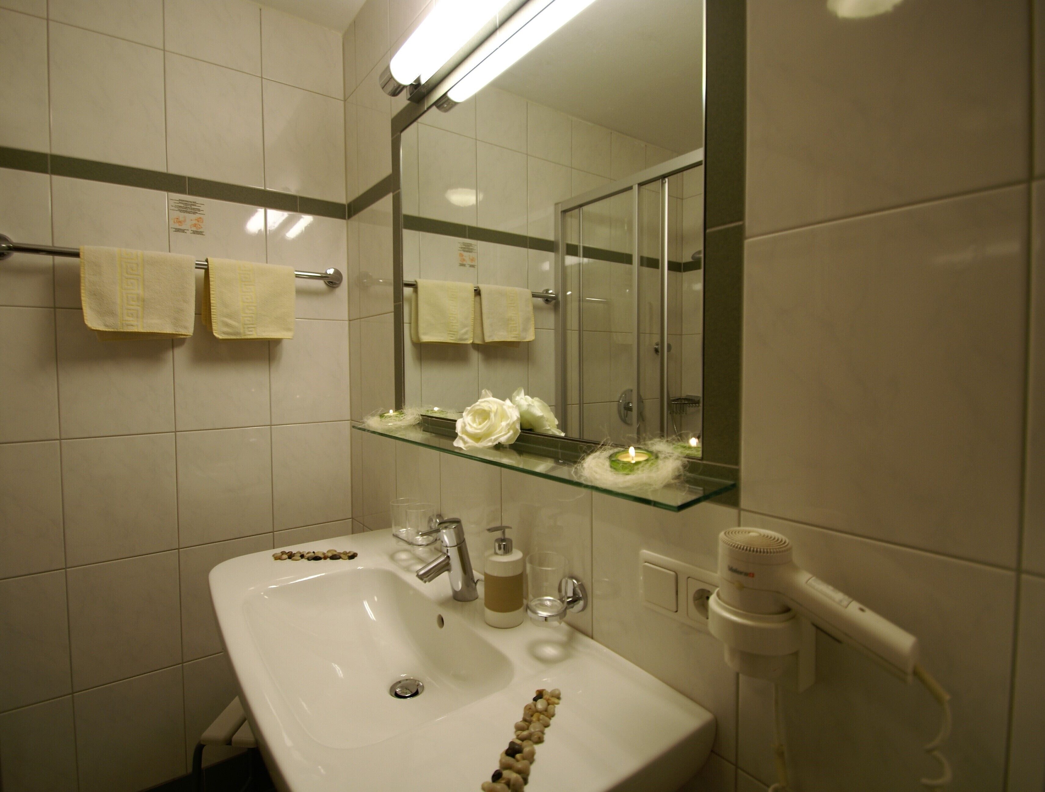 Ett badrum med ett vitt handfat, en glashylla med en vit ros och en spegel ovanför handfatet.
