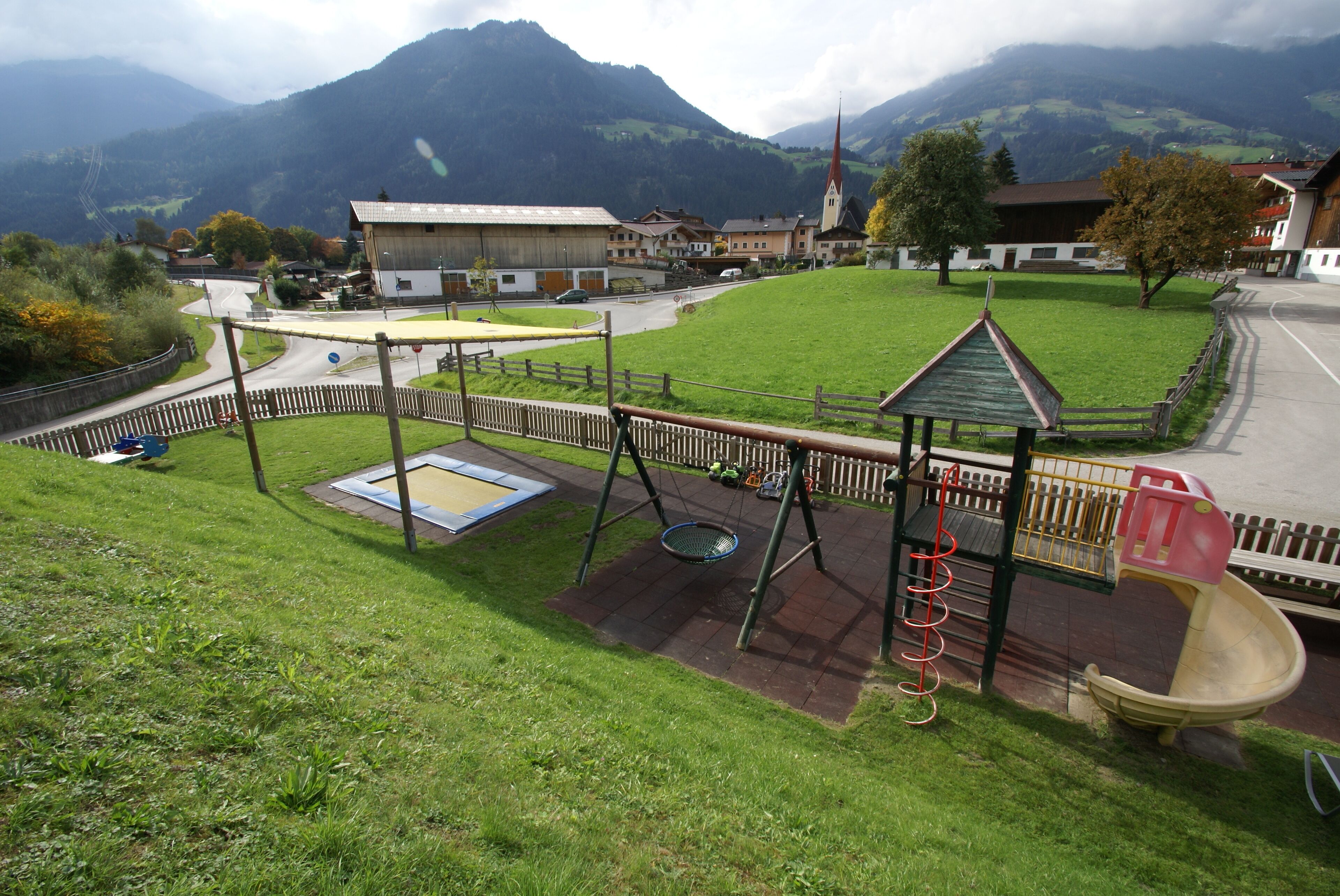 A playground with a slide and swings, surrounded by a fence, with a mountain backdrop.