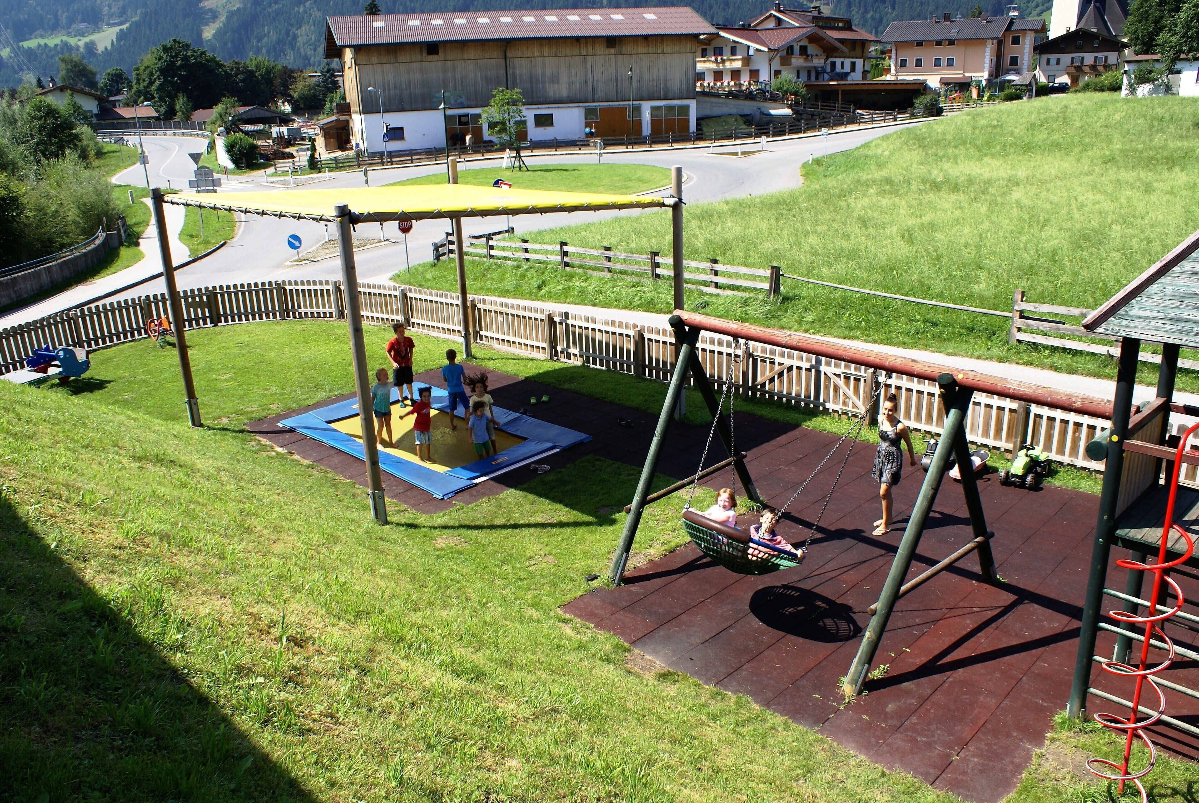 A playground with swings and a trampoline, surrounded by a wooden fence and a grassy area.