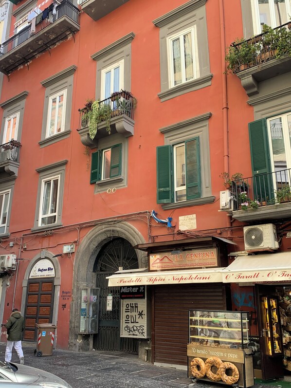 A building with multiple windows, some with shutters, and balconies with plants. There is a storefront with a sign and a display of bread.