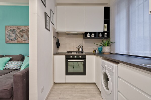 A compact kitchen with white cabinets, a stove, an oven, and a washing machine. There is a teal wall with a world map and framed pictures.