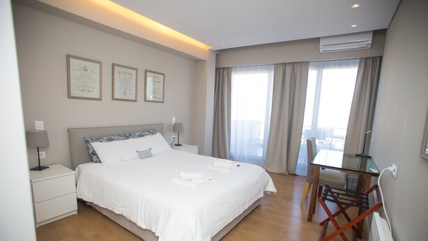 Quarto de hotel com uma cama grande, mesas de cabeceira, uma escrivaninha e uma cadeira. Há cortinas nas janelas e quadros pendurados na parede.