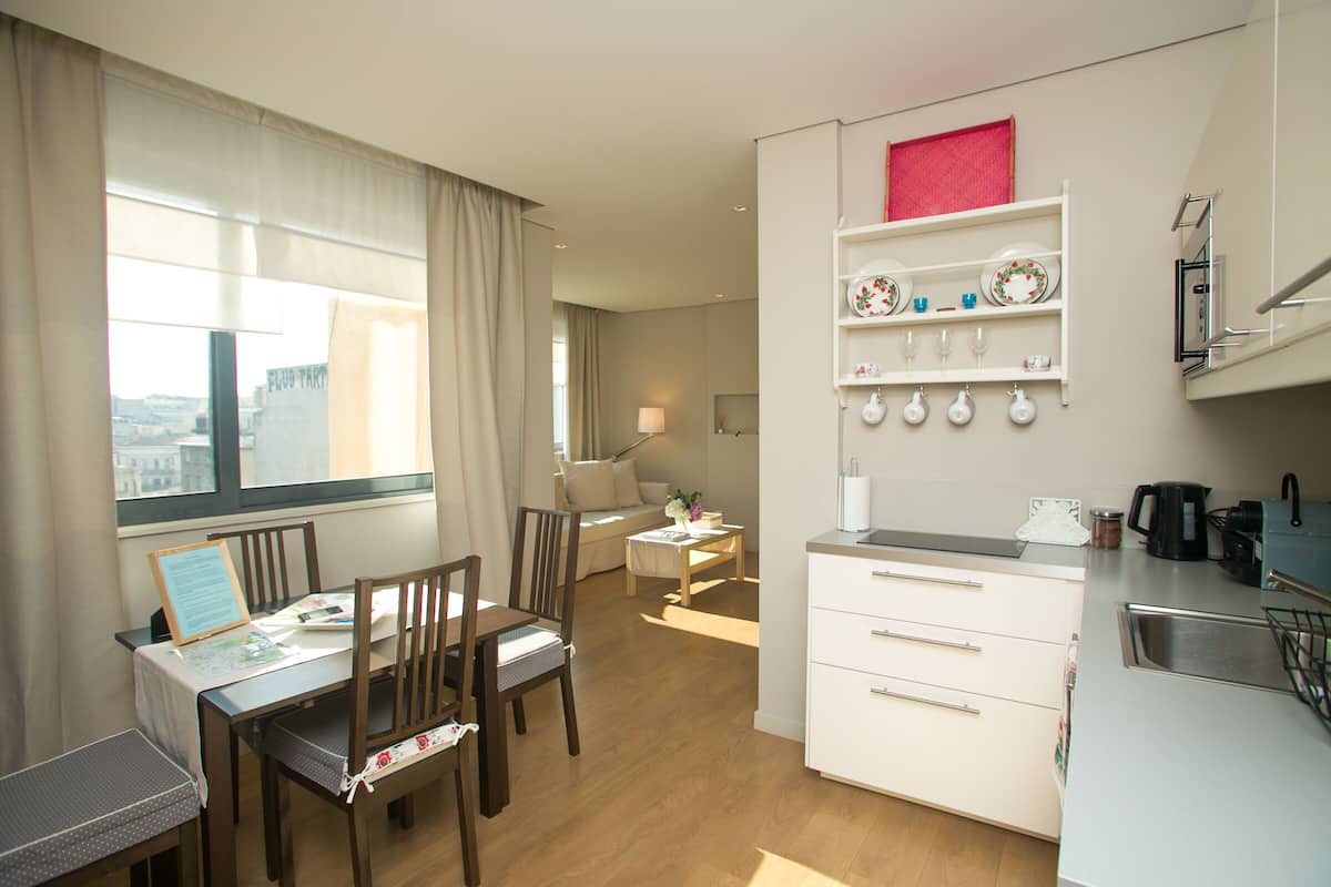 A modern kitchen with white cabinets, a dining area with wooden chairs, and a view of the city through large windows.