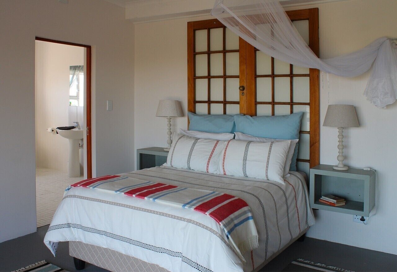 Un dormitorio con una cama, dos mesitas de noche, una mesilla auxiliar, una ventana con marco de madera y una puerta.