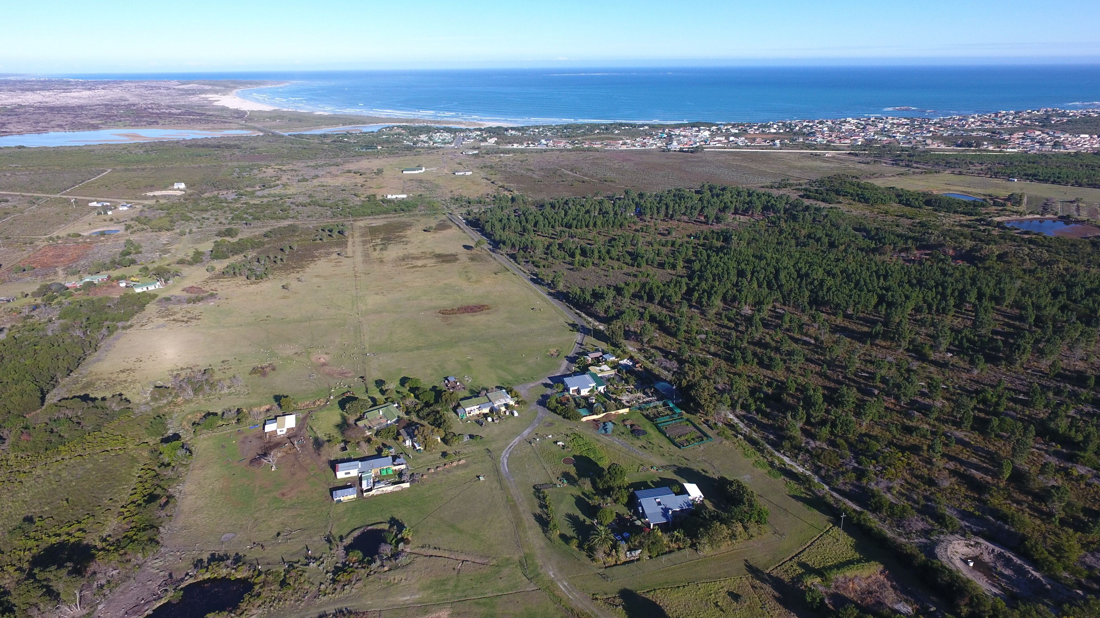 Vista aérea de una zona costera con edificios residenciales, campos abiertos y un bosque denso.