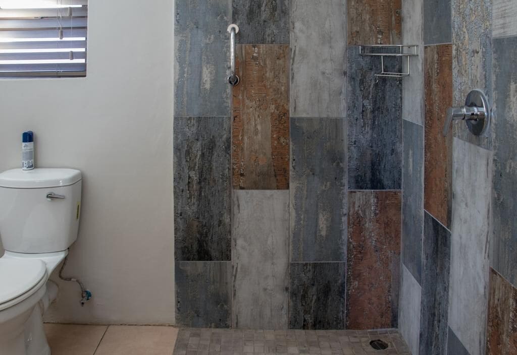 Un baño con inodoro, ducha con manija de metal y ventana con persianas.