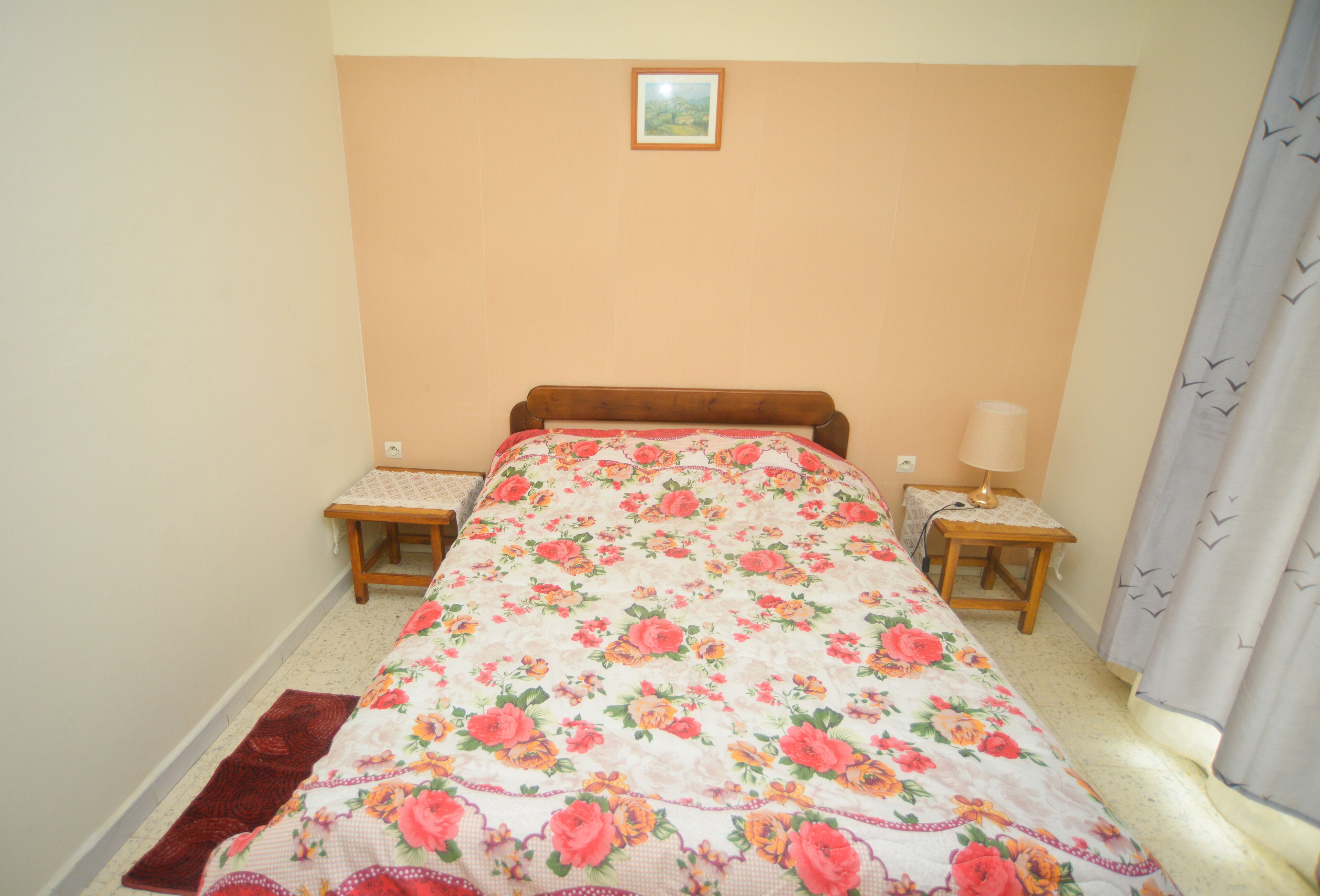 Ein Schlafzimmer mit einem blumigen Bettbezug, zwei Nachttischen, einem Beistelltisch, einer Lampe, einem gerahmten Bild an der Wand und einem Vorhang.