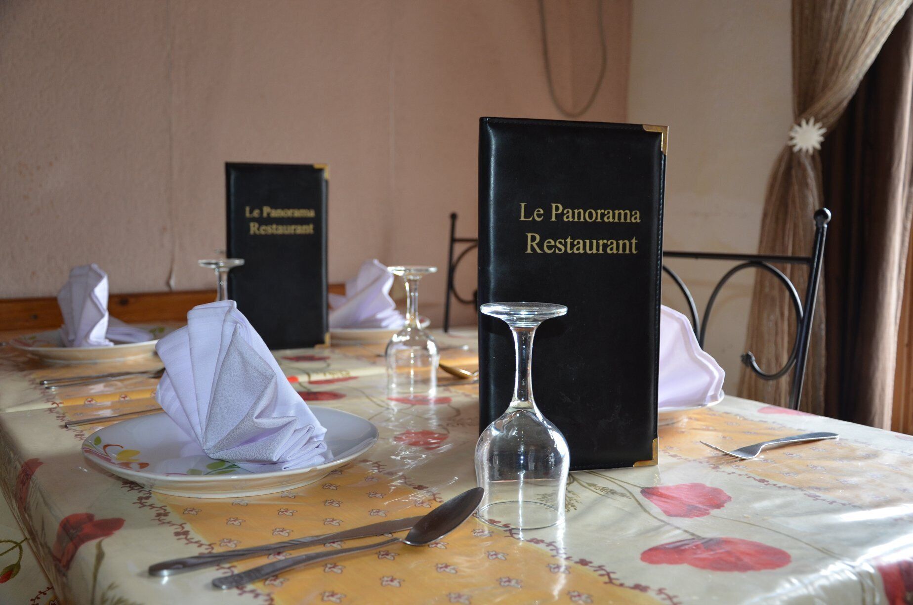 Une table de restaurant dressée avec une nappe à motifs floraux, des assiettes, des couverts et un présentoir à menus portant l’inscription « Le Panorama Restaurant ».