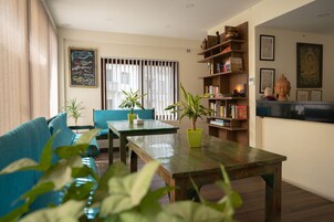 Una sala de estar con un sofá, una mesa, plantas en macetas, una estantería y una ventana con persianas.