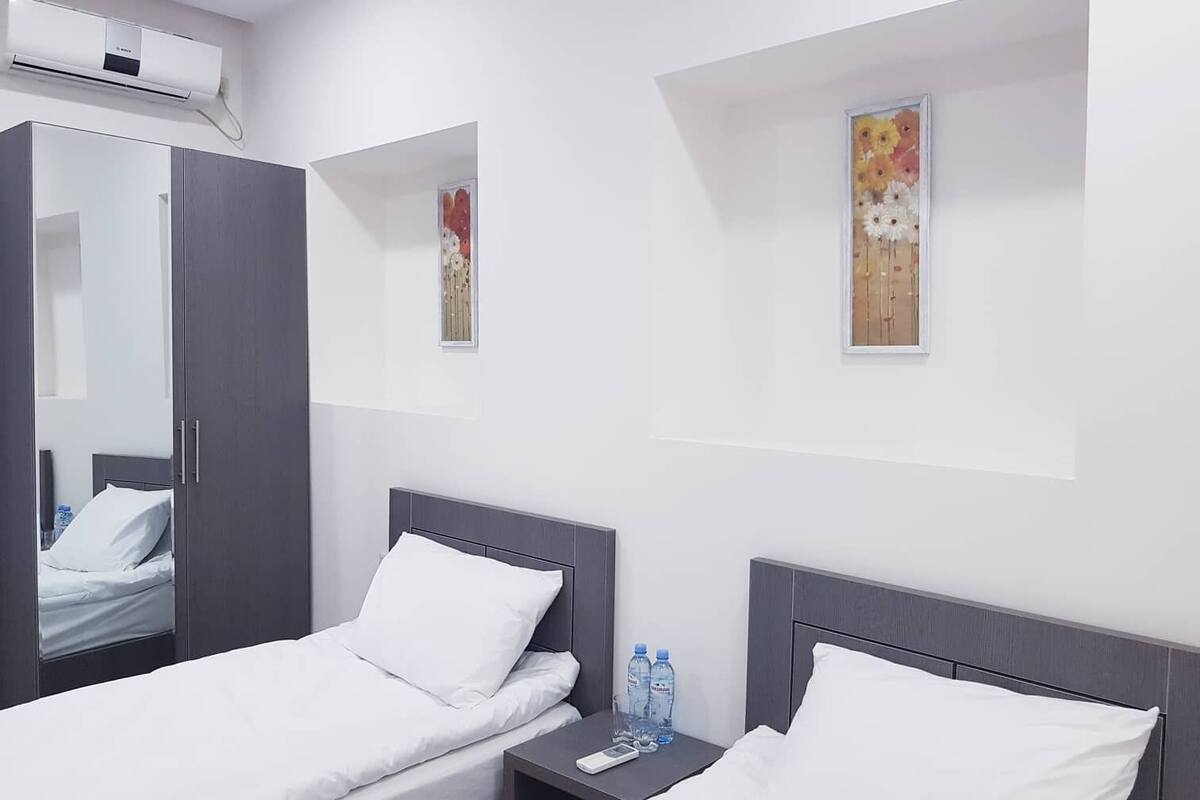 A hotel room with two beds, a wardrobe, a small window with a floral painting, and an air conditioning unit.