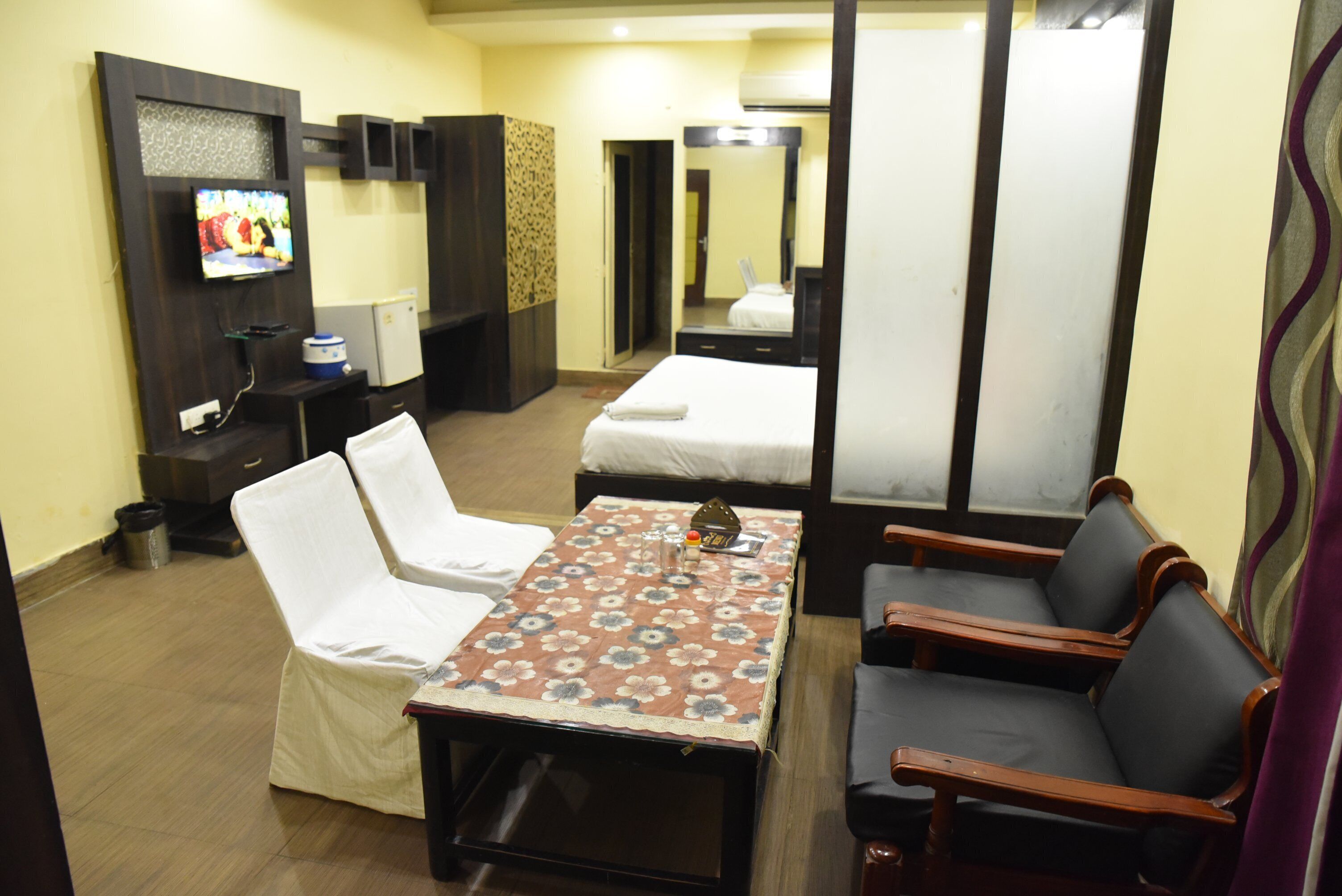 A hotel room with a bed, a small table with a floral tablecloth, two chairs, a TV on the wall, and a mirror.