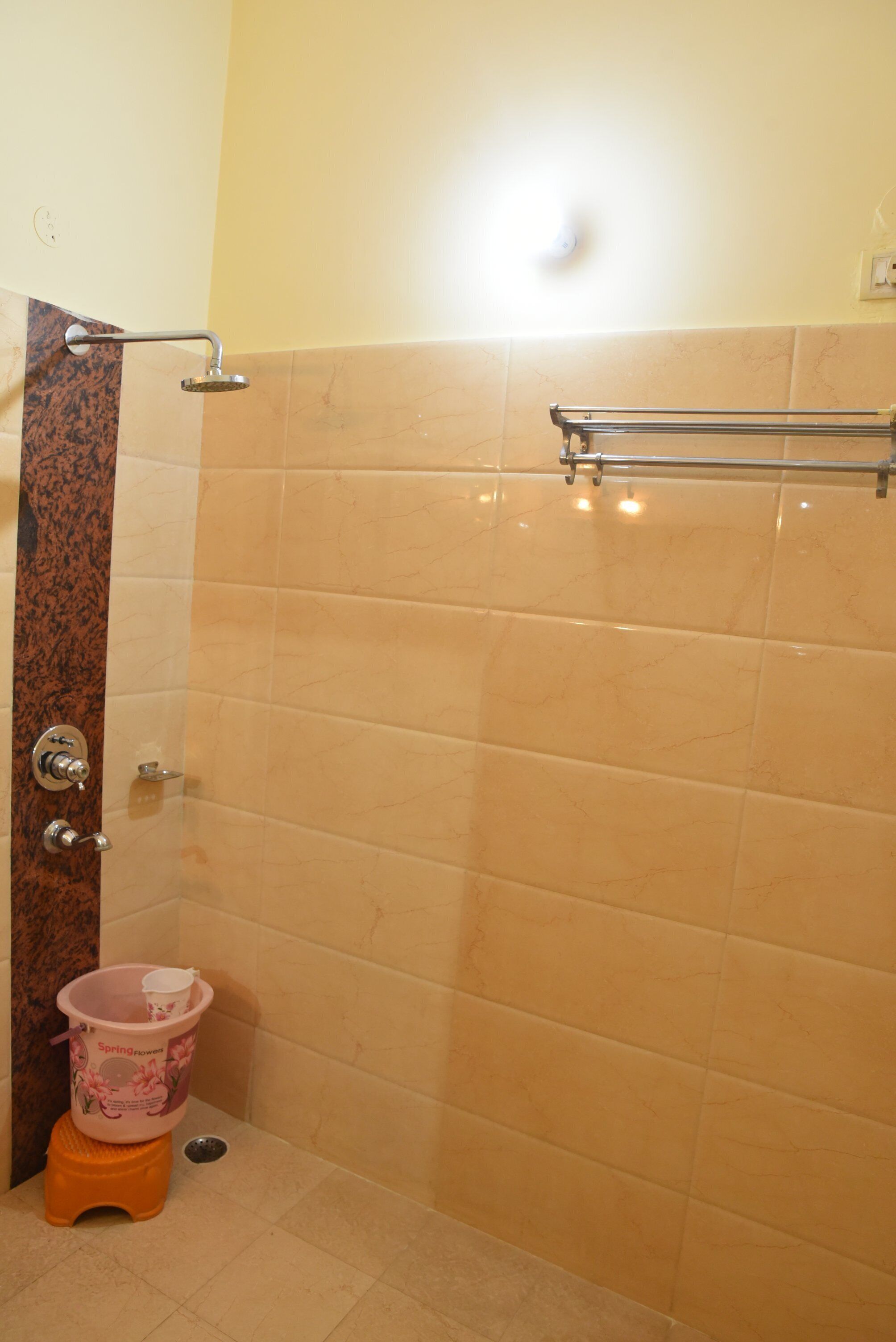 A bathroom with beige tiles, a shower with a glass door, a towel rack, and a small orange stool with a pink bucket.