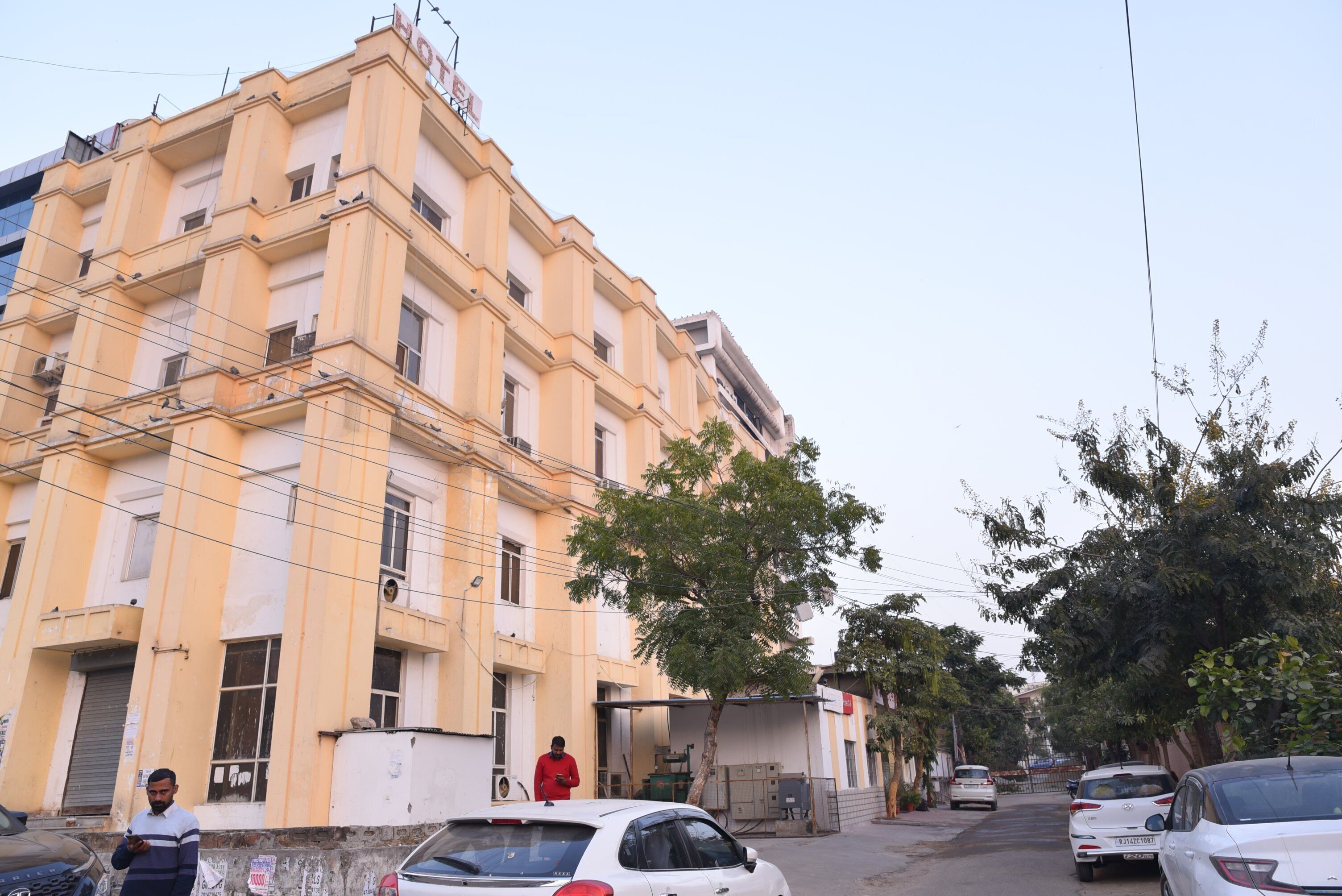 A multi-story building with a yellow facade, a tree in front, and parked cars along the street.