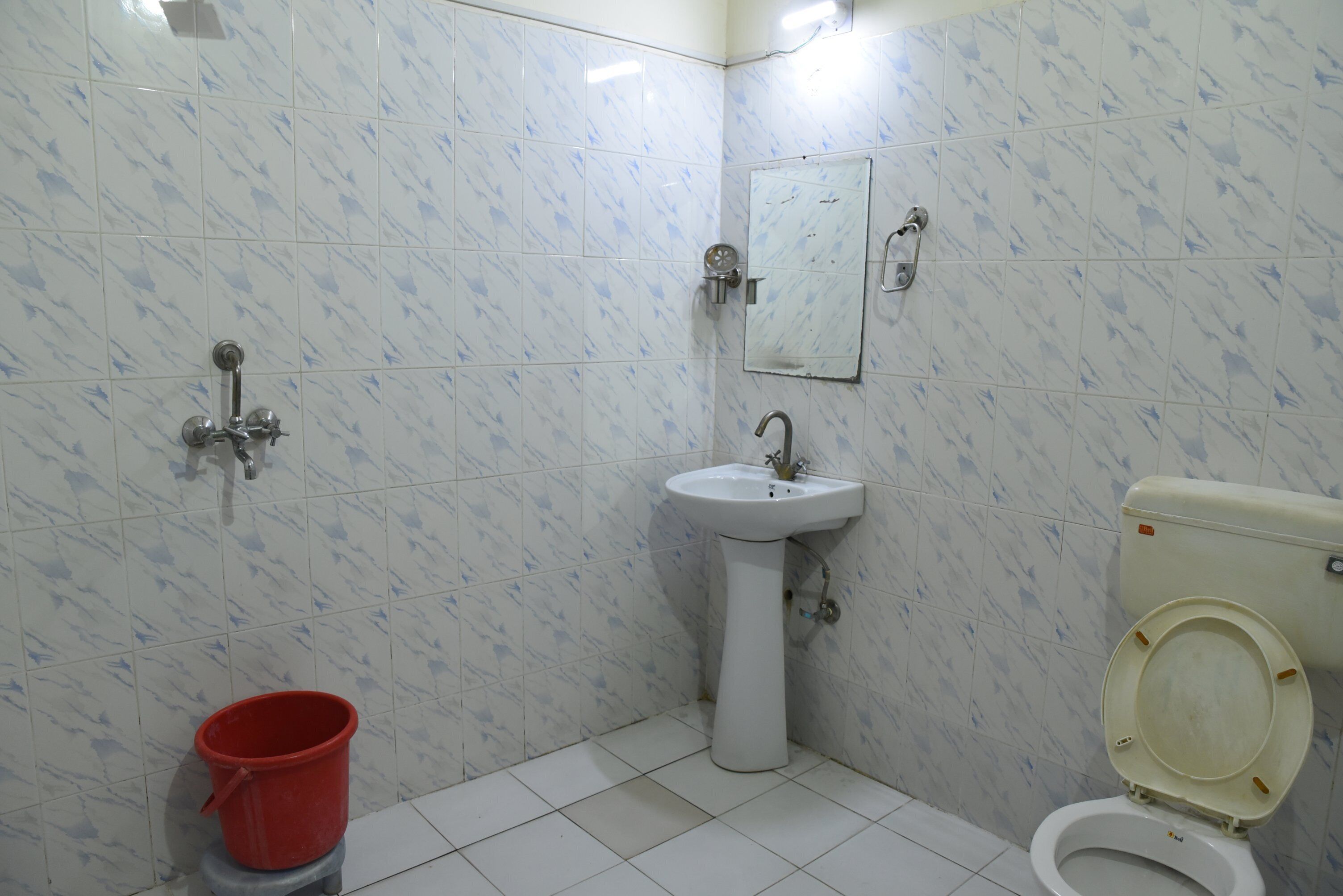 A bathroom with white tiles, a sink, a toilet, a red bucket, and a mirror on the wall.