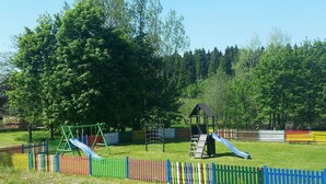 A playground with swings, slides, and a climbing structure, surrounded by a colorful fence and trees. - Landhotel Wettin (Hermsdorf-Erzgebirge)