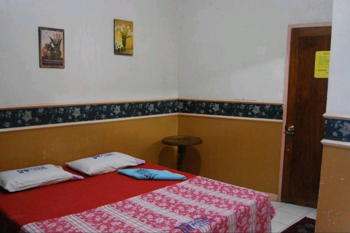 A small room with a single bed, two pillows, a small stool, and two framed pictures on the wall.